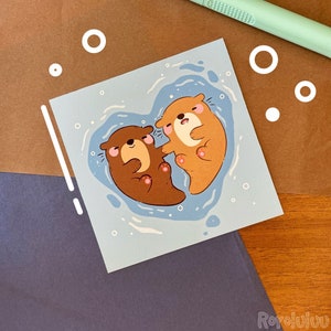 以下が含まれることがあります： ハートの形をした2匹のオッターが抱き合っている青と白のイラスト。オッターは茶色で、鼻はピンク色です。背景は薄い青色で、白い点が付いています。