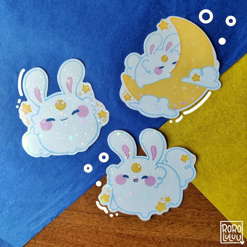 May include: Three holographic stickers featuring white rabbits with pink cheeks and a yellow crescent moon symbol on their foreheads. One sticker shows a rabbit resting on a yellow crescent moon with clouds and stars. The RORO LULUU logo is visible.