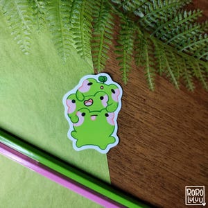 May include: A sticker featuring four green frogs stacked on top of each other. The frogs have pink cheeks and white eyes. The sticker is on a green and brown surface.