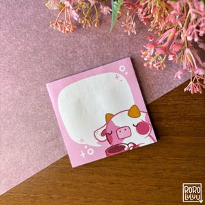 Puede incluir: Un bloc de notas adhesivas rosa con una vaca de dibujos animados sosteniendo una taza de café. La vaca tiene la nariz rosa y una mancha blanca en la frente. La nota tiene un globo de diálogo con la palabra "to" escrita dentro.