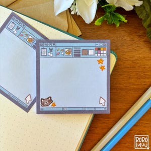 May include: Square sticky notes with a light blue computer interface design. The notes feature a cartoon raccoon, stars, and a cursor. The design includes a toolbar with icons and a colour palette. The notes are on a wooden surface with a notebook and pencils.