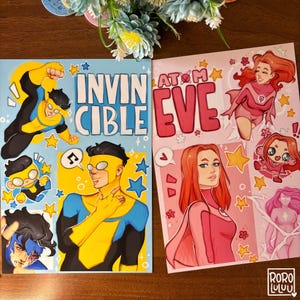 May include: Two comic book prints featuring superhero characters. One print has the text "INVINCIBLE" with illustrations of a yellow and blue costumed character. The other print has the text "AT M EVE" with illustrations of a pink costumed character.