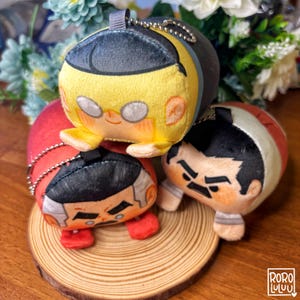 May include: Three plush character keychains in yellow, red, and white, each with a unique facial expression. The keychains are small, cylindrical, and have a silver chain for attachment. They are arranged on a small wooden slice.