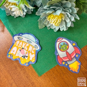 Op de afbeelding: Twee kleurrijke stickers met ruimte-thema ontwerpen. Eén toont een UFO met de tekst "TAKE ME AWAY" en een koe. De andere toont een raket met een alien erin. Beide stickers hebben een blauwe rand en glitteraccenten.