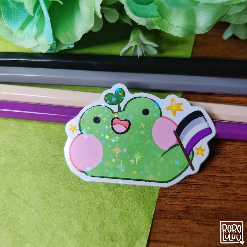 May include: A green frog with a pink belly and a small plant on its head holds a black, white, and gray flag. The frog is surrounded by glitter and stars.
