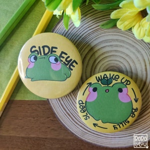 Könnte beinhalten: Zwei gelbe Anstecknadeln mit Frosch-Illustrationen. Die erste Nadel zeigt einen grünen Frosch mit rosa Wangen, der seitlich schaut und den Text "Side Eye". Die zweite Nadel zeigt einen grünen Frosch mit rosa Wangen und einem grünen Spross auf dem Kopf und den Text "Wake Up, Still Sleepy".