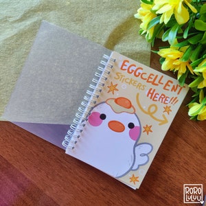 Op de afbeelding: Een klein, geel en wit spiraalnotitieblok met een cartoon-eierfiguur en de tekst "Eggcellent Stickers Here!!". Het notitieblok heeft een transparante plastic hoes.