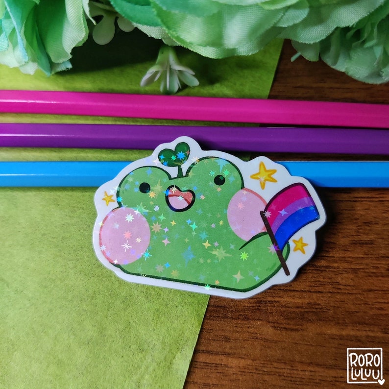 May include: A green frog sticker with a sparkly iridescent finish. The frog is holding a small bisexual pride flag. The sticker has a white outline and is on a wooden surface.