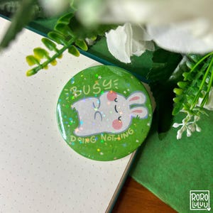 May include: A green button with the text "BUSY DOING NOTHING" and a cartoon rabbit illustration. The button has a holographic glitter effect. The button is set against a background of a notebook and greenery.