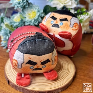 May include: Two plush character dolls with red and white outfits, black hair, and stern facial expressions. One doll is on a small wooden slice, and the other is behind it. The dolls have keychains attached.