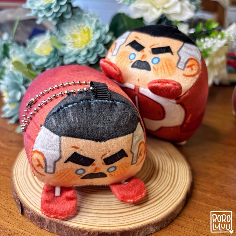 May include: Two plush character dolls with red and white outfits, black hair, and stern facial expressions. One doll is on a small wooden slice, and the other is behind it. The dolls have keychains attached.