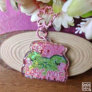 May include: A pink and green acrylic keyring with a frog design and the words "BASIC MATH". The keyring has a pink clover-shaped clasp and a silver ring. The frog has closed eyes and is decorated with hearts.