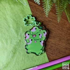 May include: A green acrylic keychain featuring a stack of three cartoon frogs with pink accents. The frogs are smiling and have their tongues sticking out. The keychain is attached to a green clover-shaped charm.