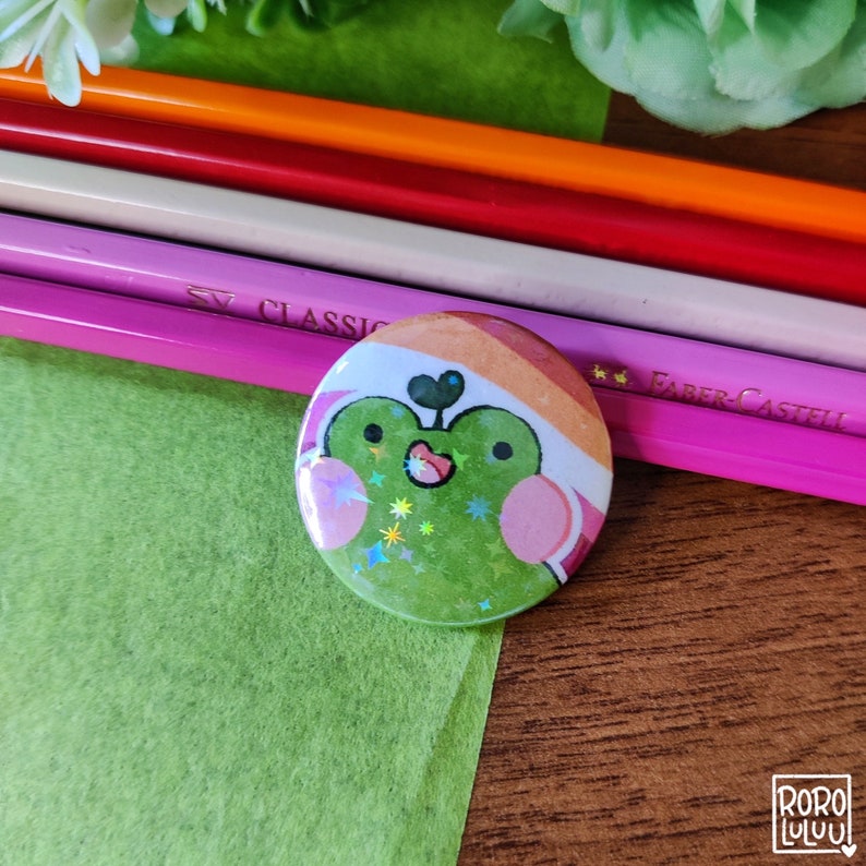 May include: A green frog wearing a pink and orange hat with glitter and stars. The frog is smiling and has pink cheeks. The frog is on a button.