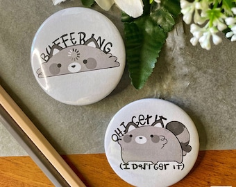 May include: Two round, white button pins with a cartoon raccoon design. The first pin says "Buffering" and the second pin says "Oh I get it (I don't get it)".