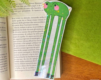 Könnte beinhalten: Ein hohes, grünes, holografisches Lesezeichen mit einem Cartoon-Frosch mit langen Beinen und rosa Wangen. Der Frosch hat einen kleinen, neutralen Gesichtsausdruck und trägt schwarze Stiefel an den Enden seiner verlängerten Beine.