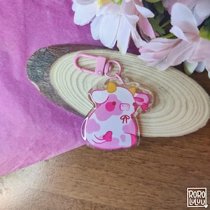 以下が含まれることがあります： ピンクと白の牛のキーホルダーで、ピンクのリボンが付いています。キーホルダーは透明なアクリル製で、金属製のリングが付いています。