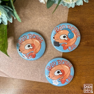 Può includere: Tre bottoni rotondi azzurri del Bad Posture Club. Ogni bottone presenta un gamberetto dei cartoni animati con la scritta "Bad Posture Club". Un gamberetto indossa le cuffie, un altro usa un computer portatile e il terzo legge.