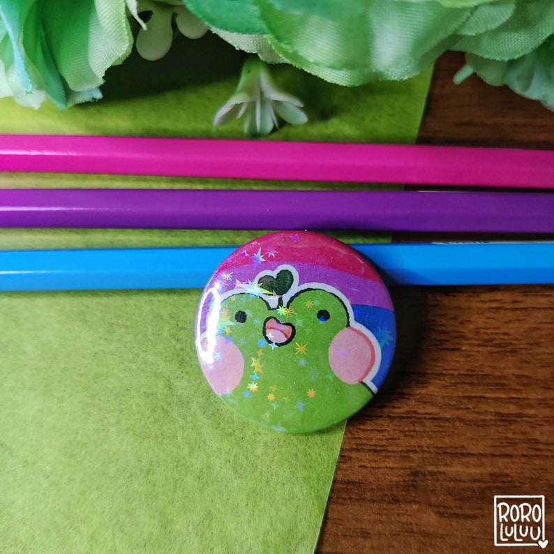 May include: A green frog with a pink heart and a rainbow flag on its back. The frog is surrounded by glitter and stars. The frog is on a button.