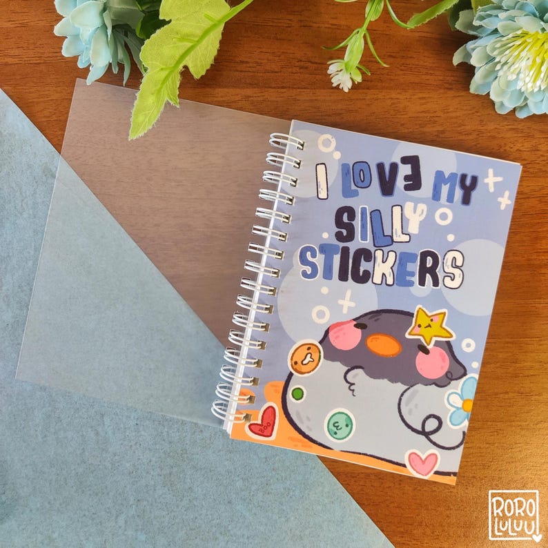 May include: A blue and white spiral bound notebook with the text "I love my silly stickers" and a cartoon penguin with stickers on it.