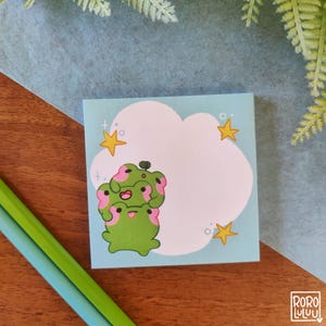 Könnte beinhalten: Ein blauer und weißer Notizblock mit einer Illustration einer rosa und grünen Frosch. Der Frosch ist auf sich selbst gestapelt und hat eine Wolke und Sterne im Hintergrund.