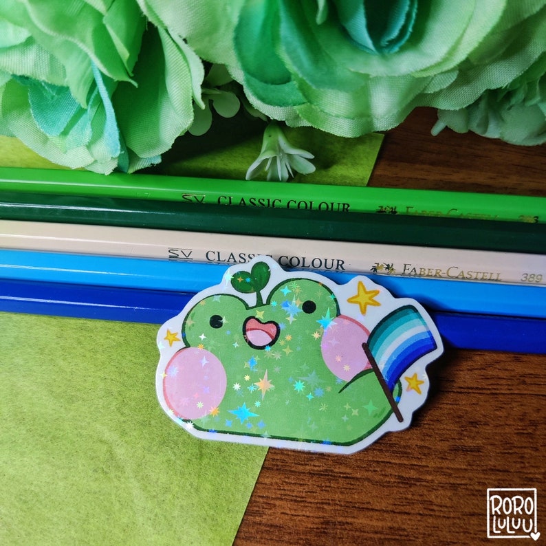 May include: A green frog sticker with a pink heart, a rainbow flag, and glitter. The sticker is on a wooden surface with green pencils.