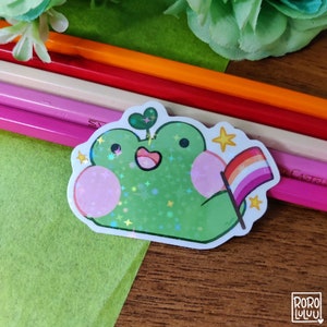 May include: A green frog sticker with a pink heart, a plant growing from its head, and a rainbow pride flag. The frog is holding the flag with its right hand and has a star on its left hand. The sticker has a holographic finish.