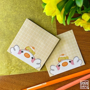 May include: Two yellow gridded sticky note pads with a cartoon illustration of a white chick with a yellow chick on top of its head. The chicks are peeking over the top of the note pads.
