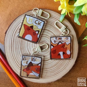 Könnte beinhalten: Drei quadratische Acryl-Schlüsselanhänger mit Cartoon-Fuchs-Illustrationen. Einer zeigt einen Fuchs mit dem Text HECK, ein anderer mit dem Text I TAKE BAD DECISIONS und der dritte einen Fuchs mit einem Messer und dem Text KEEP TESTING ME.