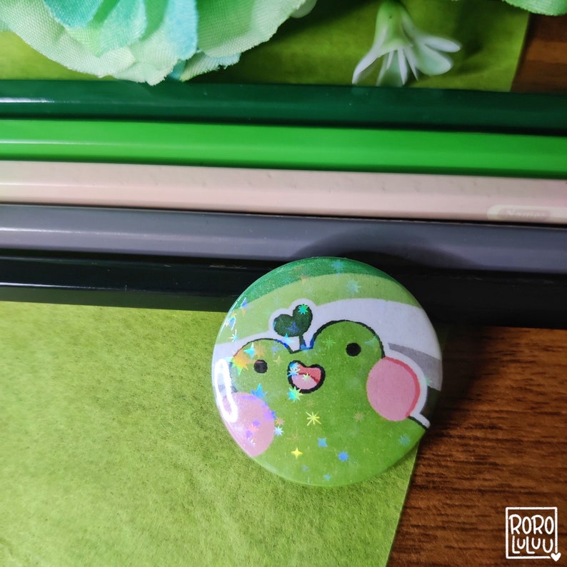 May include: A green and white button with a cartoon frog wearing a green clover on its head. The frog has a pink nose and cheeks. The button has a glittery finish and a white background with green stripes.