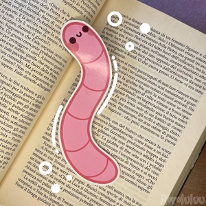 以下が含まれることがあります： 笑顔、黒い目、ピンクの頬を持つ、S字型のピンク色のミミズのステッカー。ステッカーには白い縁取りがあり、イタリア語のテキストが印刷された開いた本の上に置かれています。デザインはシンプルで、体の節が線で描かれています。