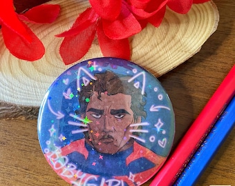 May include: A round button with a glittery blue background and a cartoon illustration of a person with brown hair and a serious expression. The person is wearing a red and blue shirt. The text "BABY GIRL" is written in pink and purple letters around the bottom of the button.