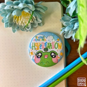 May include: A round button featuring a cartoon frog with large eyes and pink cheeks. The button has a blue background with white clouds and the text "Thinking about my hyperfixation" in rainbow colours.