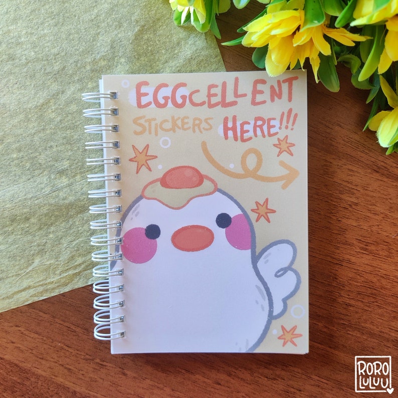 Op de afbeelding: Een spiraalgebonden notitieboek met een gele achtergrond en een cartoonkuiken met een gebakken ei op zijn kop. De tekst op de omslag luidt "Eggcellent Stickers Here!!".