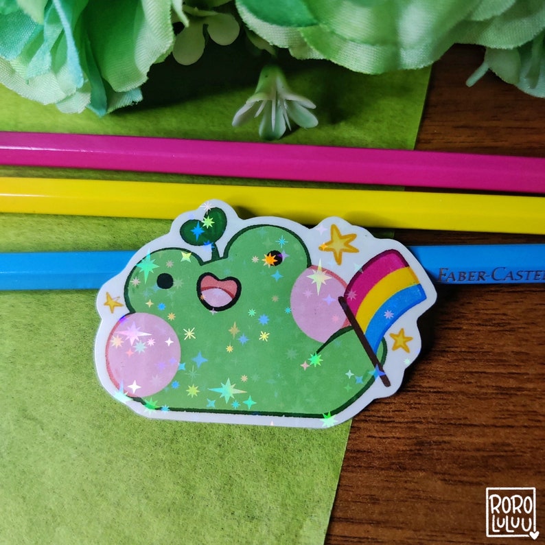 May include: A green frog with a rainbow pride flag sticker. The frog is holding the flag with its right hand. The frog is surrounded by glitter and stars.