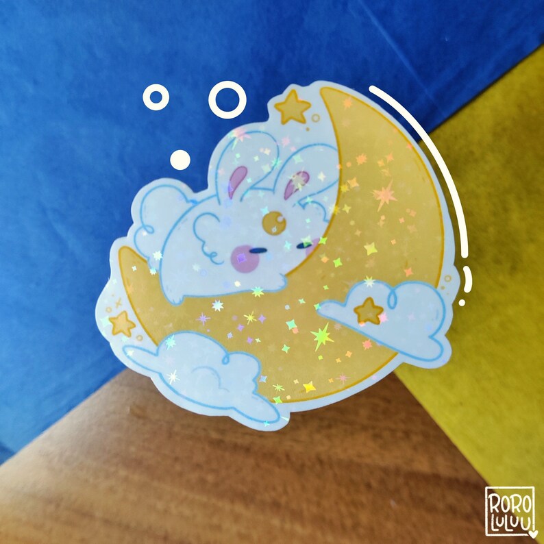 May include: A holographic sticker featuring a white rabbit with pink cheeks sleeping on a yellow crescent moon. The design includes light blue clouds, small yellow stars, and a shimmering star-patterned finish. The text RORO LULUU is visible.