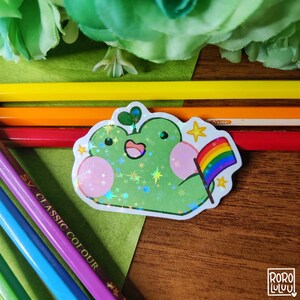 May include: A green frog with a pink belly and a rainbow flag. The frog is holding the flag in its right hand. The frog is surrounded by white stars. The background is a wooden surface.