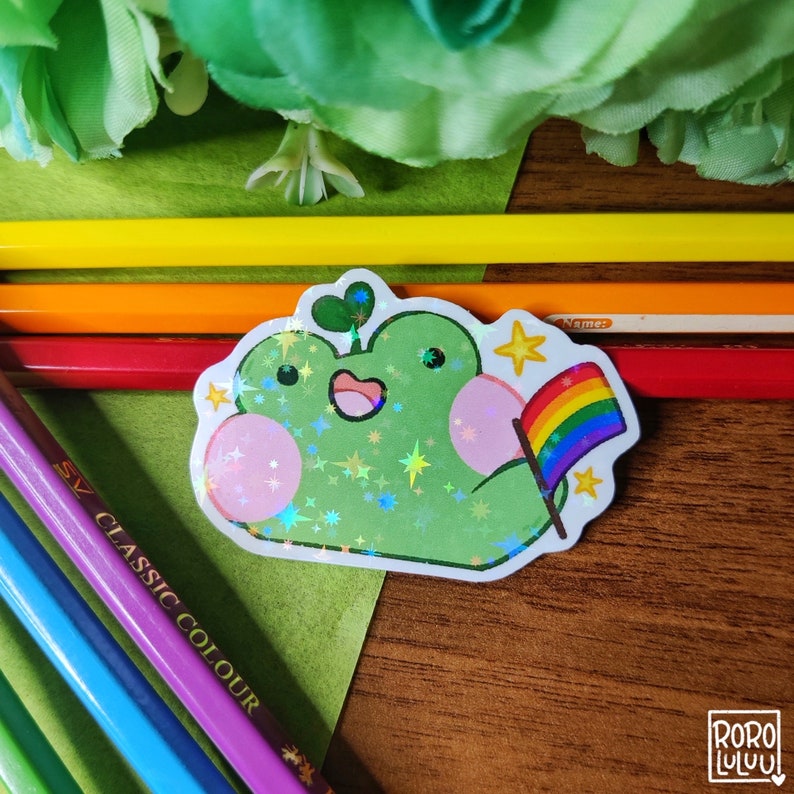 May include: A green frog with a pink belly and a rainbow flag. The frog is holding the flag in its right hand. The frog is surrounded by white stars. The background is a wooden surface.