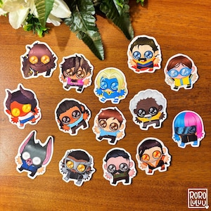 Puede incluir: Una colección de pegatinas de estilo caricatura coloridas con varios personajes con ojos grandes y atuendos únicos. Las pegatinas están dispuestas sobre una superficie de madera, con algo de vegetación en el fondo. Las pegatinas tienen un borde blanco.