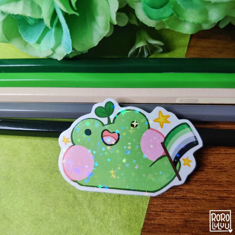 May include: A green frog sticker with a pink belly and a green, black, and white pride flag. The sticker is covered in glitter and has a holographic finish.
