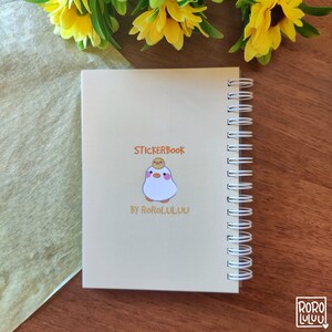 Op de afbeelding: Een cr&egrave;mekleurig spiraalnotitieblok met de tekst "STICKERBOOK BY RORO.LULUU" en een cartoonillustratie van een wit kuiken met een geel kuiken op zijn kop.