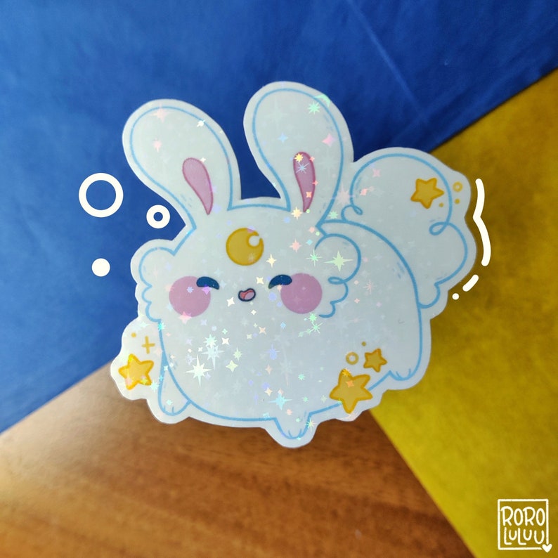 May include: A holographic sticker featuring a white, round rabbit character with long ears, pink cheeks, and a yellow crescent moon on its forehead. The design includes small yellow stars, blue outlines, and a sparkling finish with the text RORO LULUU.