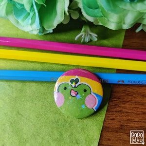 May include: A green frog with pink cheeks and a sparkly green body is wearing a pin with the pansexual pride flag colors. The pin is on a wooden surface with three colored pencils.