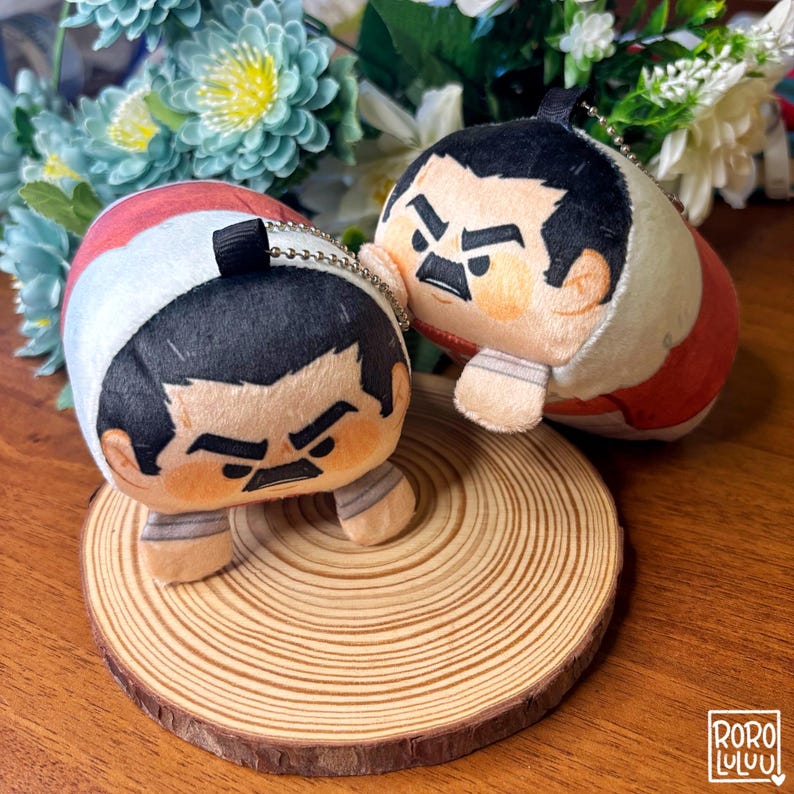 May include: Two plush sumo wrestler keychains. Each keychain features a cartoon-style wrestler with a stern expression, black hair, and a red and white outfit. They are on a wooden slice, with flowers in the background.