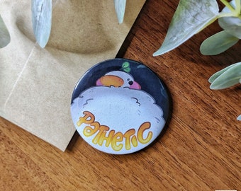 May include: A round button with a white fluffy cartoon bird with a yellow beak and a green leaf on its head. The bird is looking up and the text "PATHETIC" is written in yellow letters around the bird.