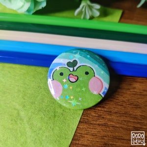 May include: A green frog with a pink nose and cheeks, wearing a blue and green striped hat with white stars, on a round button.