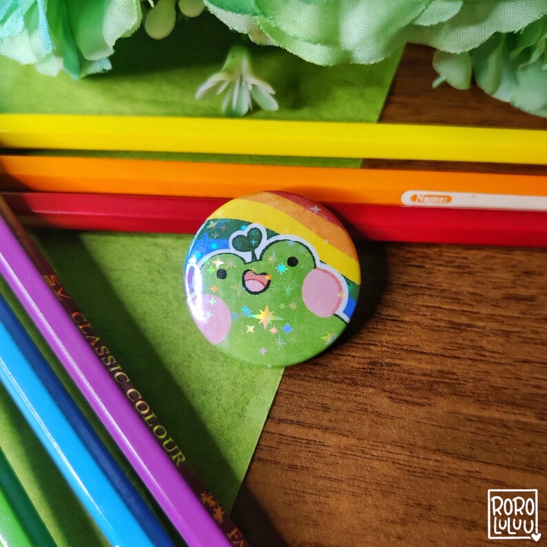 May include: A green frog with a rainbow and glitter design on a button. The button is on a wooden surface with colored pencils.