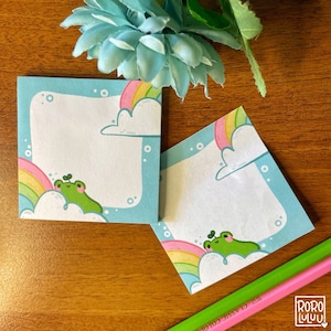 Può includere: Due blocchi per appunti quadrati con un bordo azzurro, con un ranocchio dei cartoni animati su una nuvola con un arcobaleno. I blocchi hanno un centro bianco per la scrittura. Una matita verde e rosa è in primo piano. I blocchi sono decorati con un design carino e stravagante.