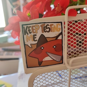 Peut inclure: Un autocollant avec un renard de dessin animé tenant un couteau et le texte "Keep Testing Me". Le renard est rouge avec des yeux blancs et se trouve sur un fond brun.