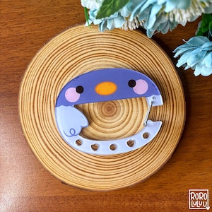Peut inclure: Un mousqueton violet et bleu clair représentant un visage de pingouin avec un bec jaune et des joues roses. La partie inférieure du mousqueton présente de petites découpes de formes variées et un ressort métallique est fixé sur le côté droit.